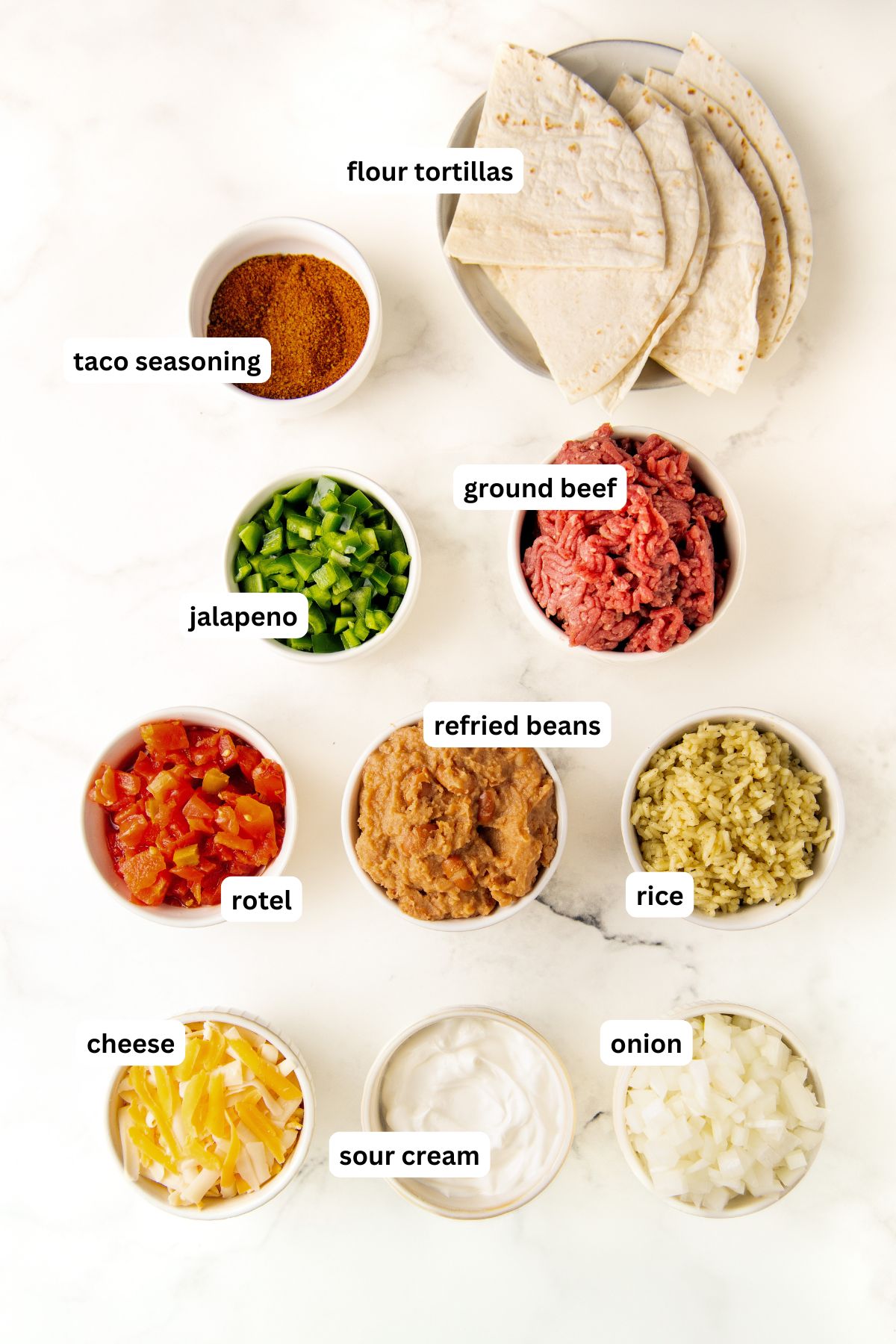 Ingredients for burrito casserole recipe in order from top to bottom: flour tortillas, taco seasoning, jalapeño, ground beef, Rotel, refried beans, rice, cheese, sour cream, and onion.