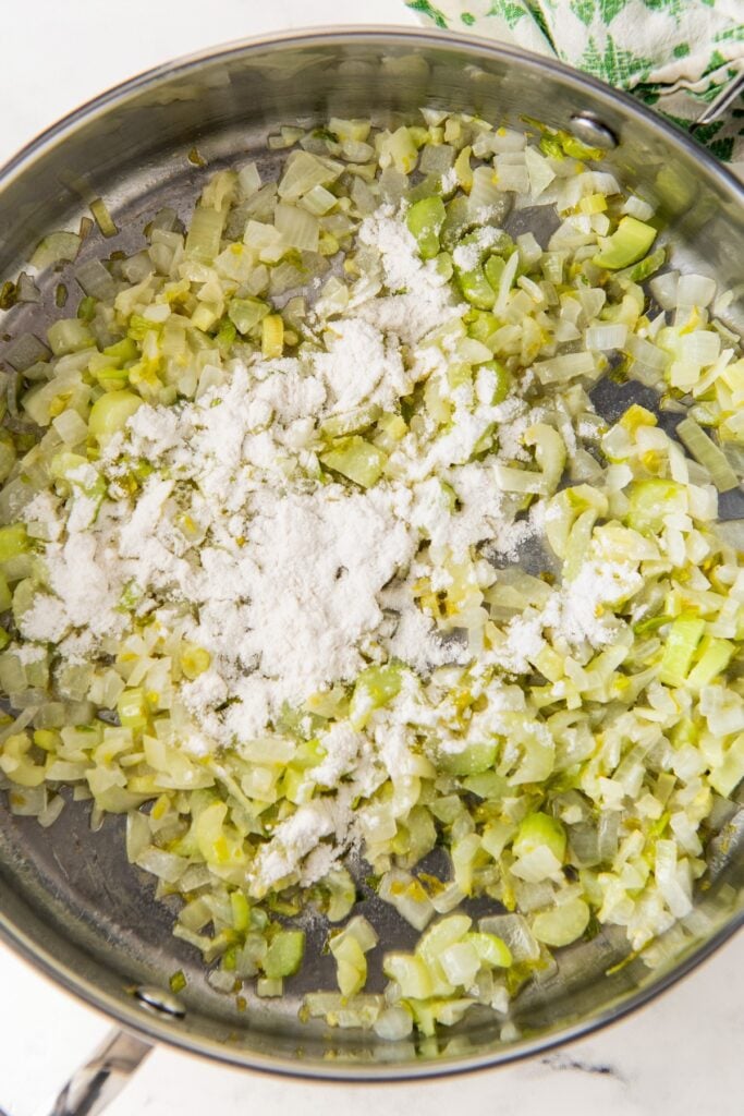 Flour is being stirred into a skillet with sautéed vegetables.