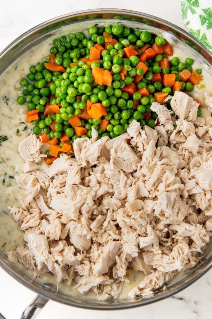 Peas, carrots, and shredded cooked chicken are being stirred into a skillet.