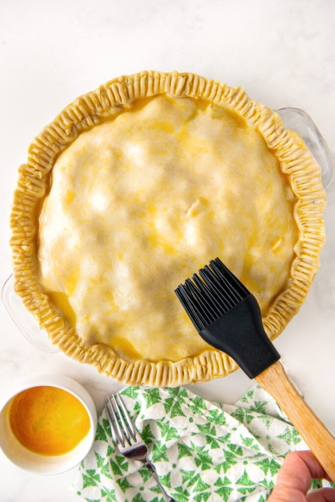 An uncooked homemade chicken pot pie is being brushed with egg wash.