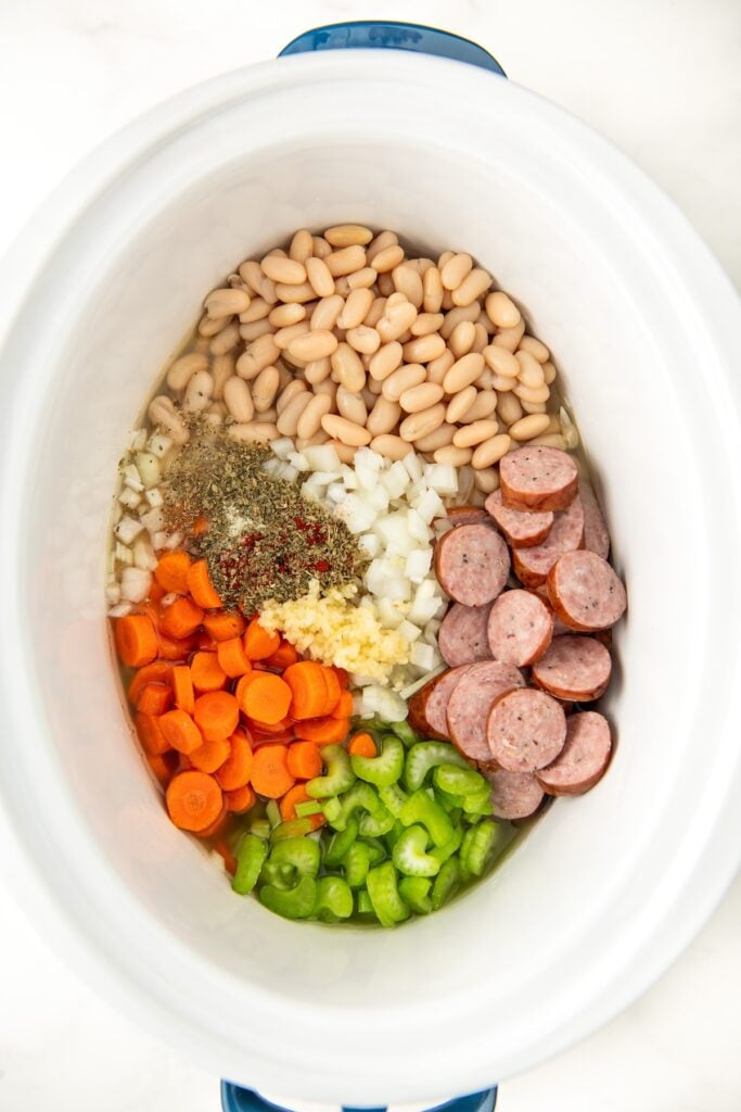 The ingredients for sausage and white bean soup are placed in a large white slow cooker.