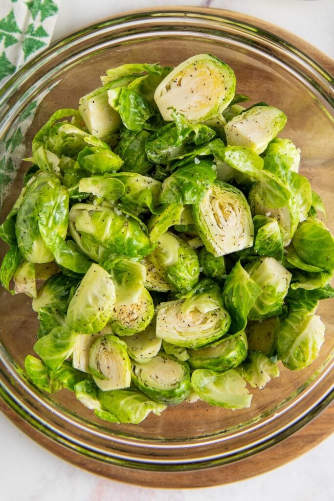 Raw, halved brussel sprouts are in a glass mixing bowl and coated with oil and seasonings.