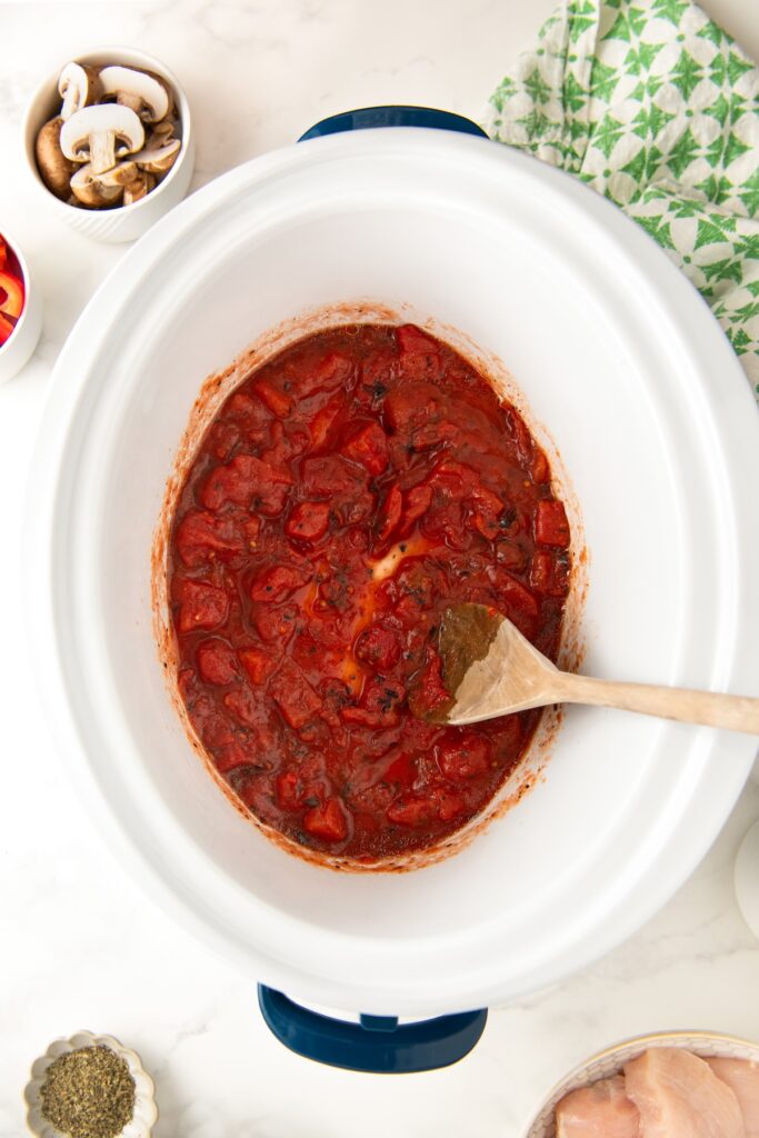 The first few ingredients for chicken cacciatore are being stirred in a white slow cooker.