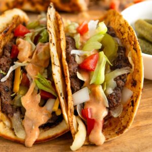 Two charred smash burger tacos filled with beef, shredded lettuce, diced tomatoes, onions, and cheese, topped with a creamy Big Mac burger sauce. The tacos are on a wooden board, with a side of pickles in a small white bowl.