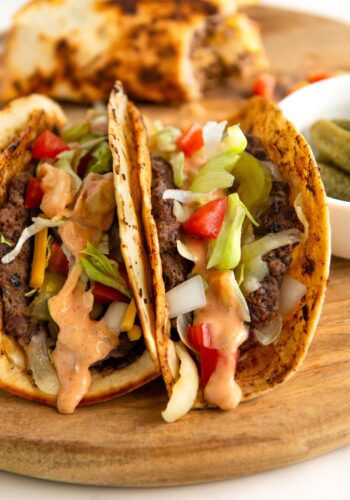 Two charred smash burger tacos filled with beef, shredded lettuce, diced tomatoes, onions, and cheese, topped with a creamy Big Mac burger sauce. The tacos are on a wooden board, with a side of pickles in a small white bowl.