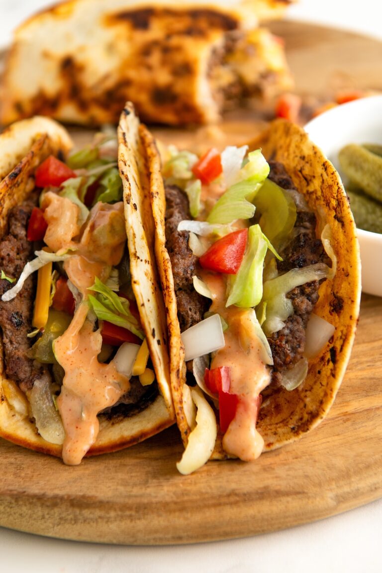 Two charred smash burger tacos filled with beef, shredded lettuce, diced tomatoes, onions, and cheese, topped with a creamy Big Mac burger sauce. The tacos are on a wooden board, with a side of pickles in a small white bowl.