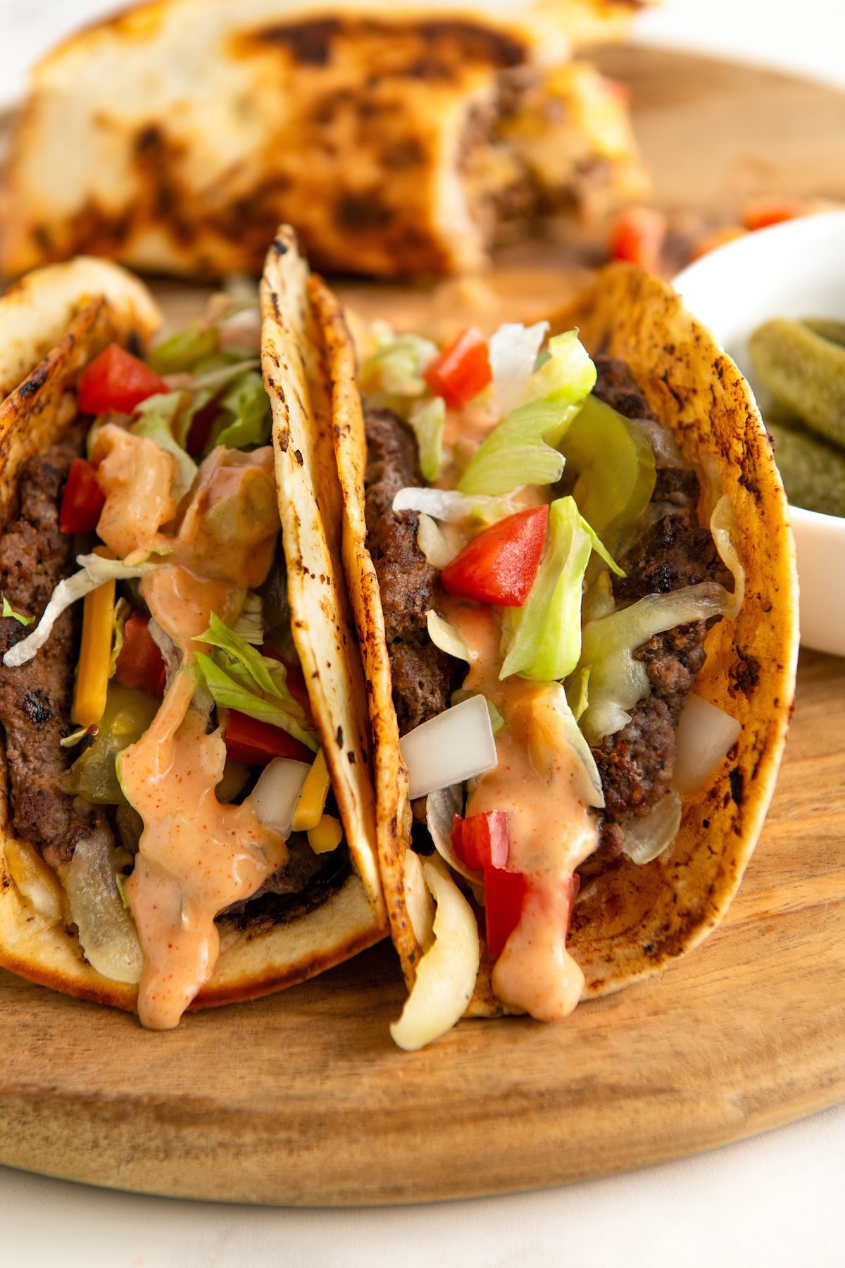 Smash Burger Tacos combine juicy burger patties with melted cheese, burger sauce, and all your favorite taco toppings in a crispy tortilla. Two charred smash burger tacos filled with beef, shredded lettuce, diced tomatoes, onions, and cheese, topped with a creamy Big Mac burger sauce. The tacos are on a wooden board, with a side of pickles in a small white bowl.