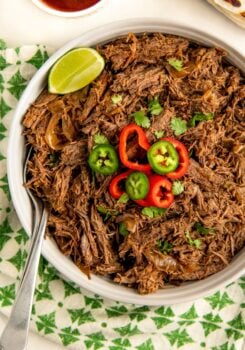 A bowl of Mexican shredded beef garnished with slices of red and green chili peppers, cilantro, and a lime wedge. A spoon rests in the bowl, which is placed on a green and white patterned cloth. A small bowl of sauce is visible in the background.