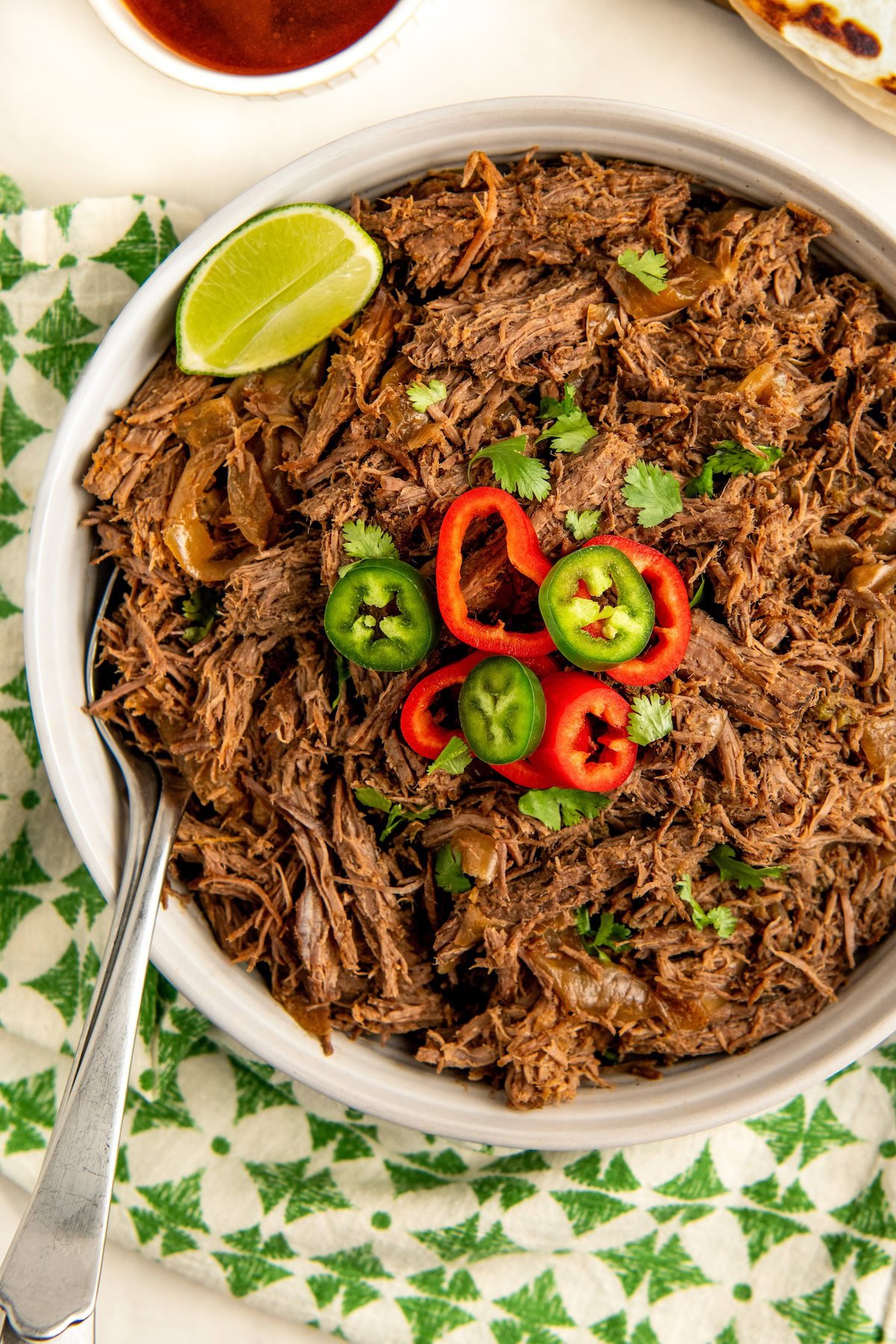 So tender and flavorful, this easy Crockpot Mexican Shredded Beef recipe is the perfect filling for tacos, burritos, rice bowls and more! A bowl of Mexican shredded beef garnished with slices of red and green chili peppers, cilantro, and a lime wedge. A spoon rests in the bowl, which is placed on a green and white patterned cloth. A small bowl of sauce is visible in the background.