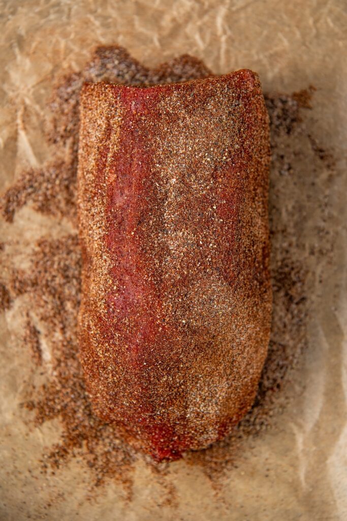 A raw beef roast covered in a brown spice rub rests on crinkled parchment paper. The seasoning appears coarse and evenly spread, creating a textured surface over the meat.