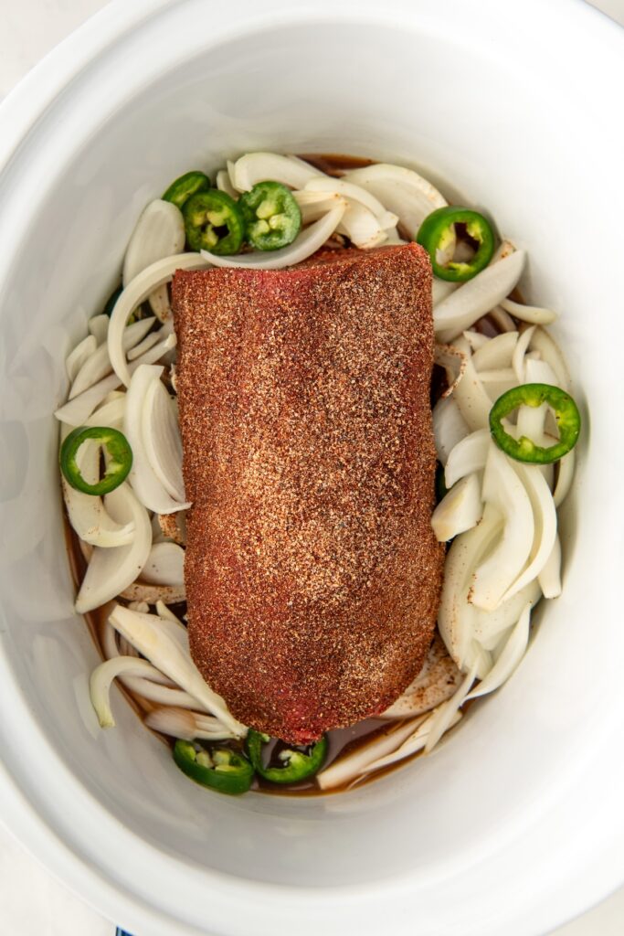 A seasoned piece of meat rests in a slow cooker atop a bed of sliced onions and jalapeños. The spices form a dark crust on the meat, and the vegetables are surrounded by a small amount of liquid at the bottom.