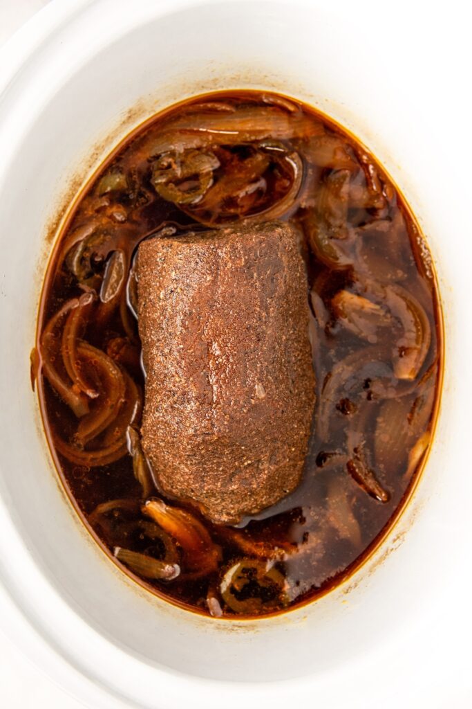 A seasoned beef roast sits in a slow cooker surrounded by cooked onions and dark, rich broth. The beef is brown with a visible spice rub, suggesting its marinated and slowly cooked to infuse flavor.