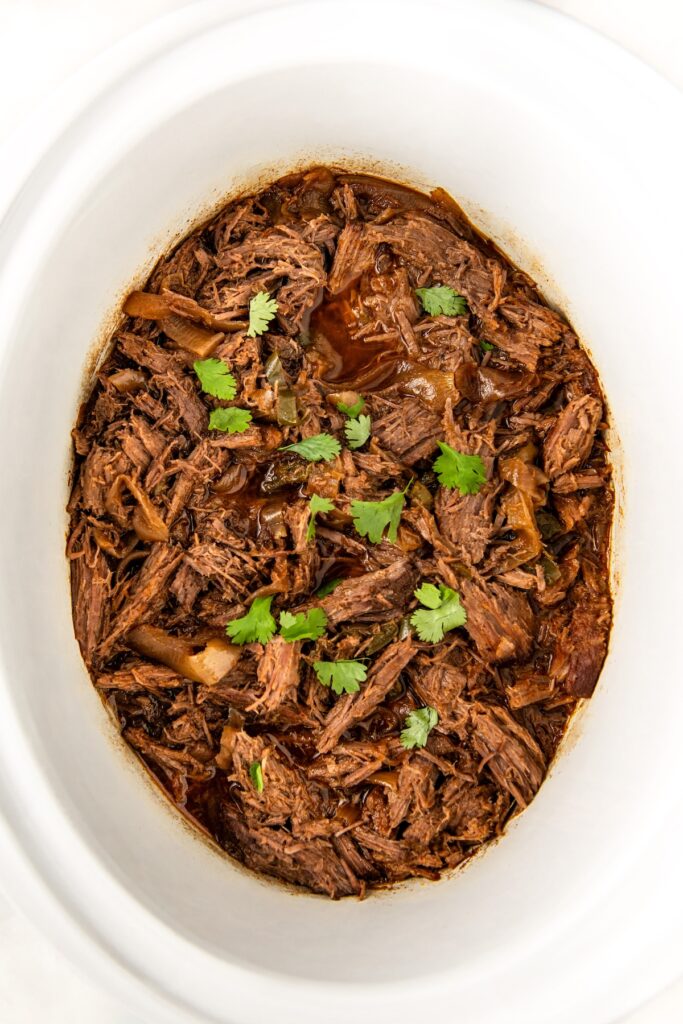 Crockpot shredded beef in a white dish, garnished with fresh cilantro. The beef appears tender, cooked in a rich, brown sauce with visible onion pieces throughout.