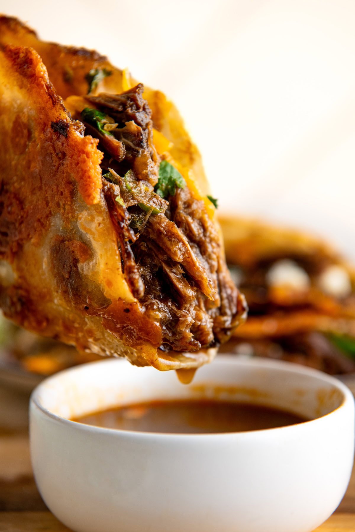 Close-up of a hand holding a cheesy Mexican shredded beef taco with tender meat and herbs, dipping it into a small white bowl of consommé. The taco is crispy and golden, with melted cheese oozing out, and the sauce is rich and slightly reddish-brown.