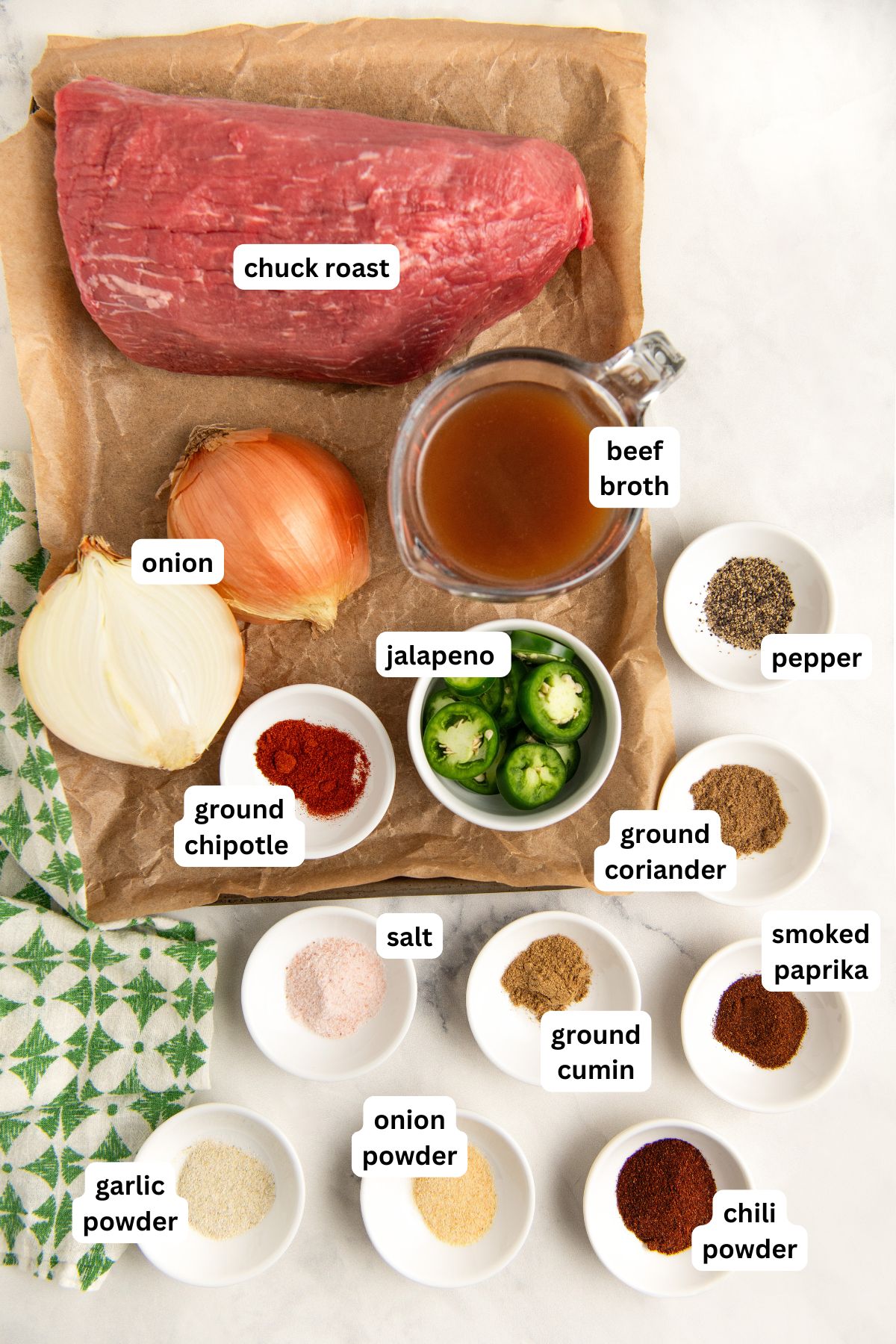 Ingredients for Crockpot Mexican Shredded Beef recipe arranged on a marble countertop: raw chuck roast on a parchment paper-lined sheet pan, two onions, a bowl of beef broth, sliced jalapeño, and spices including ground chipotle, pepper, salt, garlic powder, onion powder, ground cumin, coriander, smoked paprika, and chili powder.