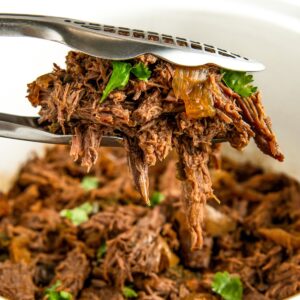 Tongs holding a serving of Crockopt shredded beef adorned with chopped cilantro, hovering above a bowl filled with more of the same beef. The meat appears tender and juicy, highlighted by small bits of onion.