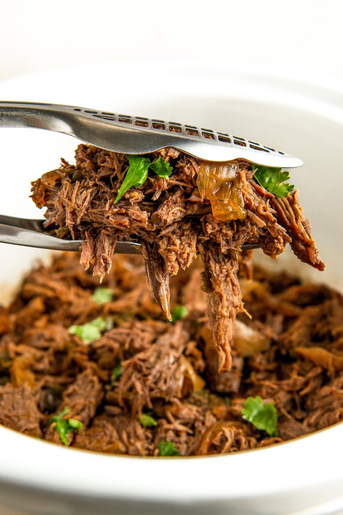Tongs holding a serving of Crockopt shredded beef adorned with chopped cilantro, hovering above a bowl filled with more of the same beef. The meat appears tender and juicy, highlighted by small bits of onion.