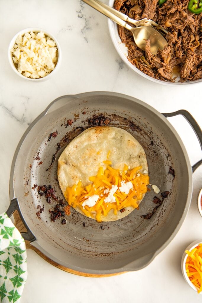 A tortilla with melted cheese and crumbled white cheese is cooking in a pan. Nearby, there are bowls with shredded meat and crumbled cheese. A green-patterned cloth is partially visible on the side.