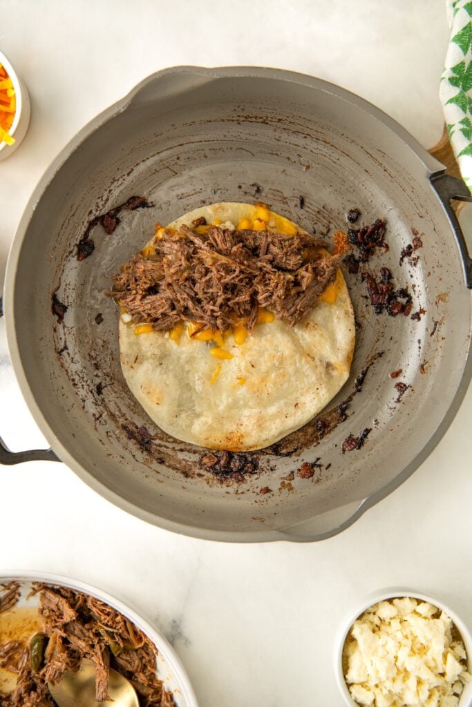 A frying pan with a tortilla filled with Mexican shredded beef and melted cheese is on a stove. The tortilla shows some browning, and the setup suggests preparation of a quesadilla. A bowl of shredded beef and a small dish of cheese are nearby.