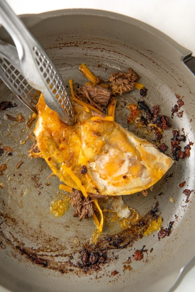 A pan with a crispy cooked, folded taco filled with melted cheese and shredded beef. A pair of metal tongs is holding the taco down into the skillet, and the pan has some browned, crispy cheese remnants.