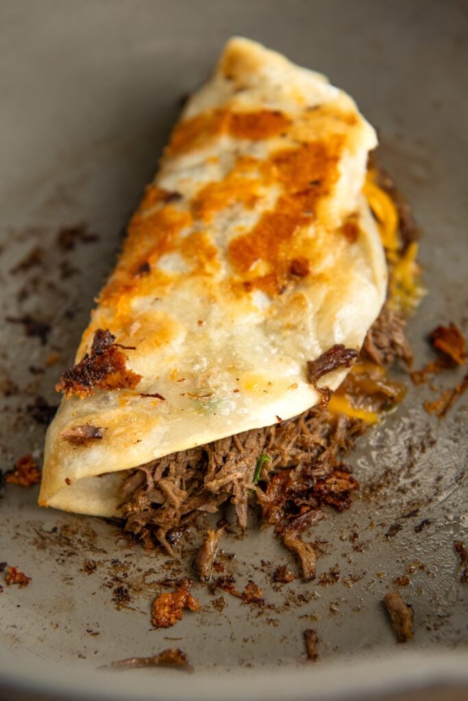 A folded taco with crispy, browned tortilla edges, filled with shredded beef and melted cheese, lying on a skillet, surrounded by leftover scattered browned meat and cheese bits.