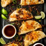 A tray of crispy shredded beef tacos filled with melted cheese and beef, garnished with jalapeño slices and lime wedges. Two small bowls of dipping sauce are beside the tacos.