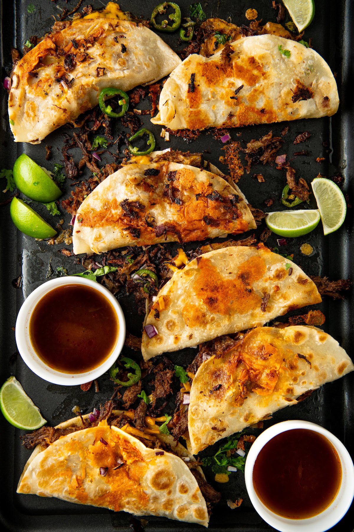 A tray of crispy shredded beef tacos filled with melted cheese and beef, garnished with jalapeño slices and lime wedges. Two small bowls of dipping sauce are beside the tacos.