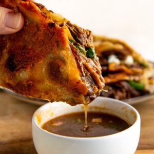 A hand dips a crispy, golden cheesy taco filled with melted cheese, shredded beef, and greens into a small white bowl of rich, brown broth. Another taco is visible in the background on a wooden surface.