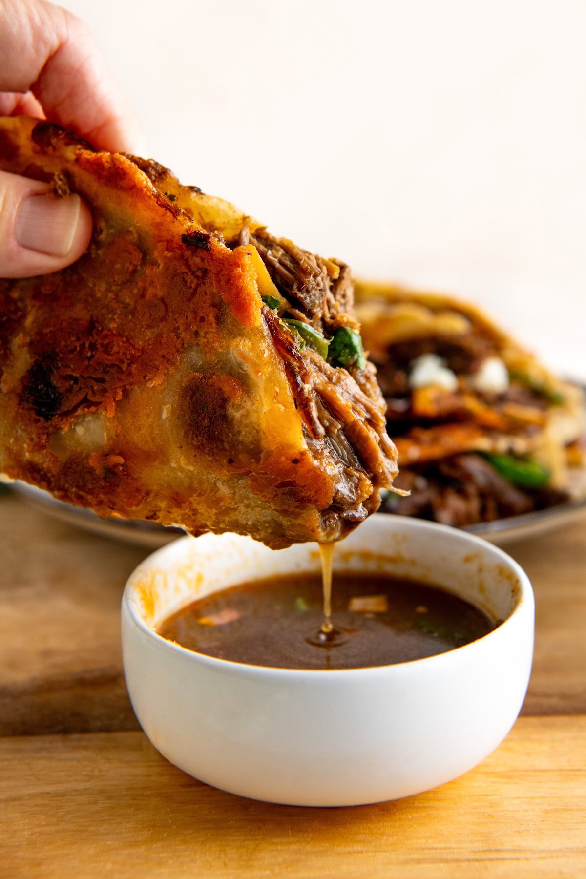 A hand dips a crispy, golden cheesy taco filled with melted cheese, shredded beef, and greens into a small white bowl of rich, brown broth. Another taco is visible in the background on a wooden surface.