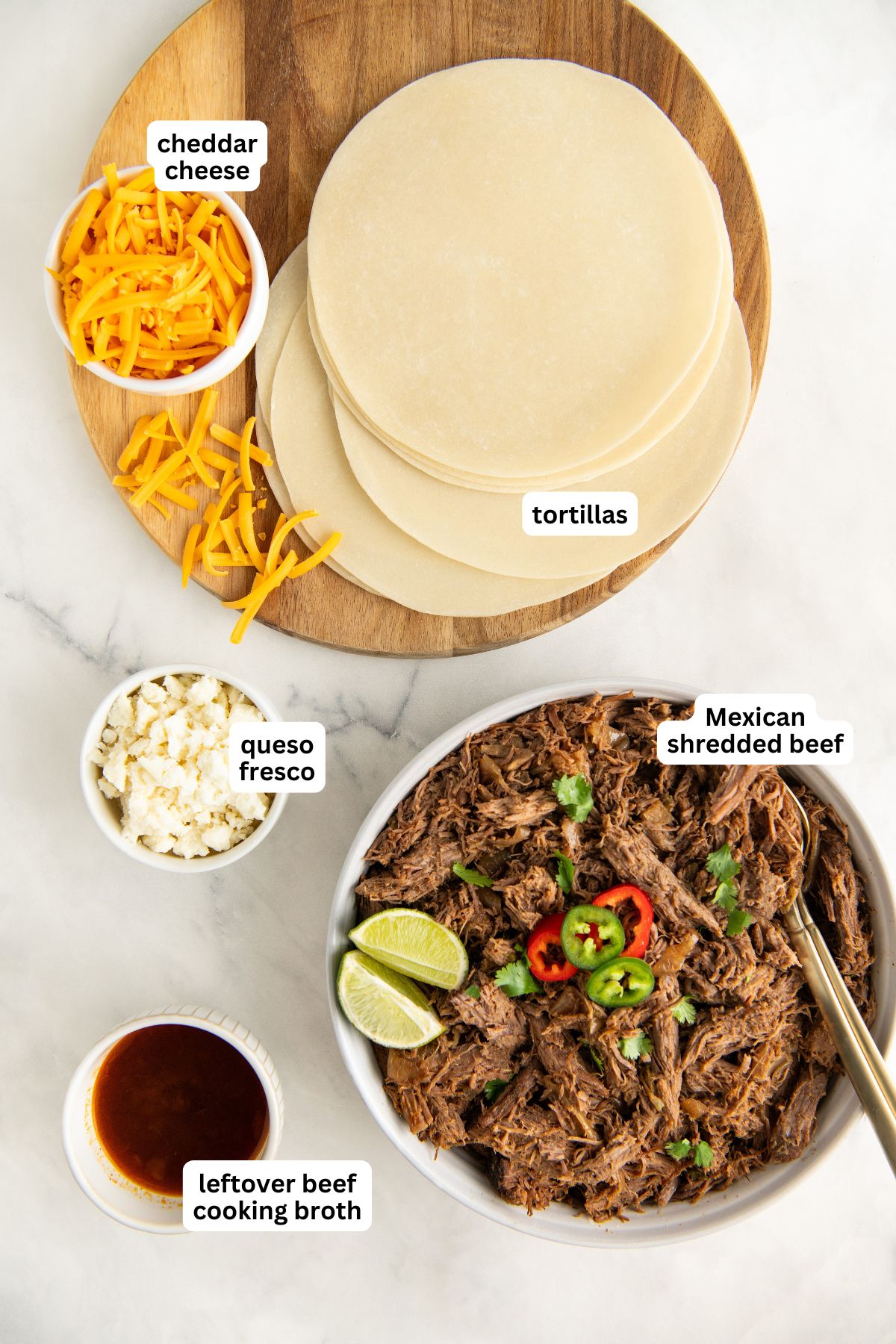 A top view of ingredients for preparing this Mexican shredded beef tacos recipe. There are tortillas, a bowl of cheddar cheese, queso fresco, Mexican shredded beef with cilantro and lime slices, and a bowl of leftover beef cooking broth.