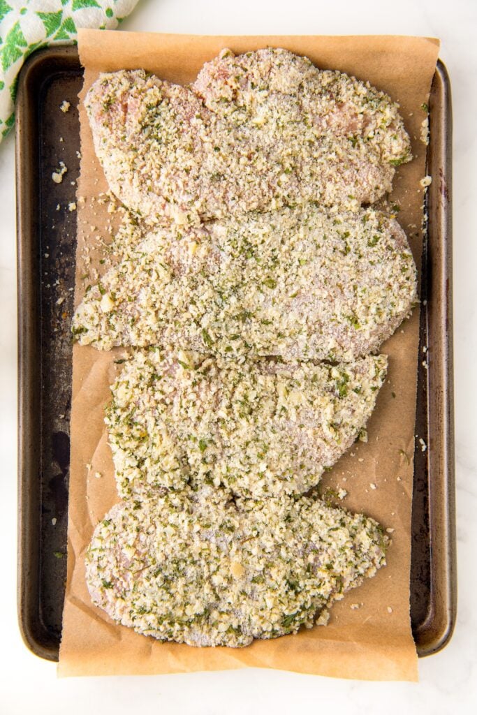 Uncooked parmesan crusted chicken breasts lined up on a prepared baking sheet.