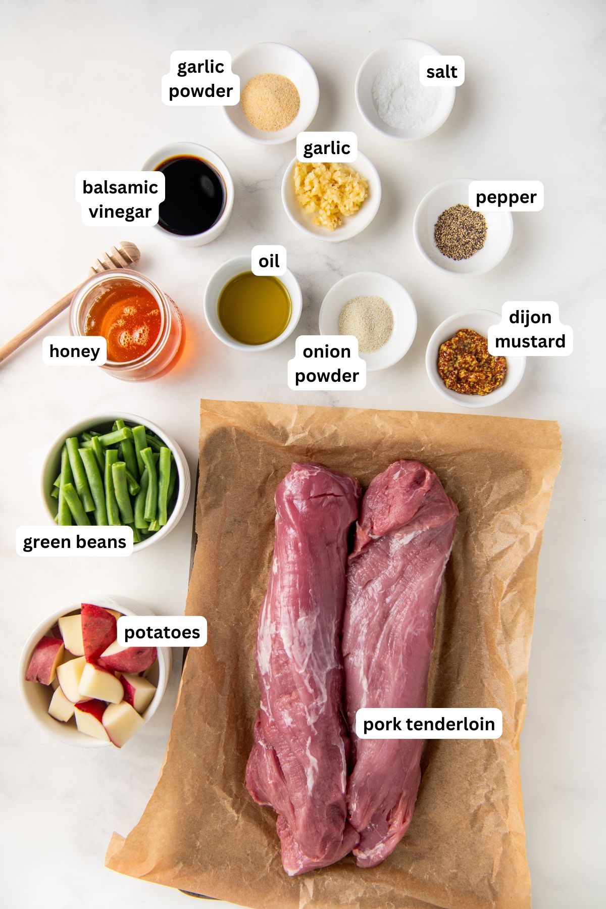 Top-down view of ingredients for honey garlic roasted pork tenderloin recipe on a white surface, labeled: pork tenderloin on parchment, sliced potatoes, green beans, garlic powder, balsamic vinegar, honey, oil, minced garlic, onion powder, salt, pepper, and Dijon mustard.