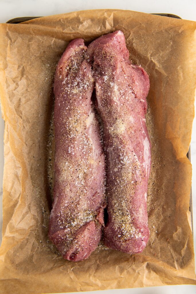 Two raw pork tenderloins seasoned with salt, pepper, garlic powder and onion powder on a parchment-lined baking sheet.