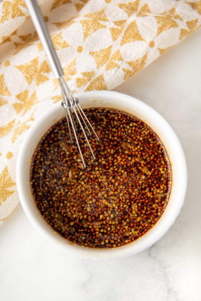 A small whisk in a white bowl filled with a dark yellow dijon mustard mixture. The bowl rests on a marble surface.