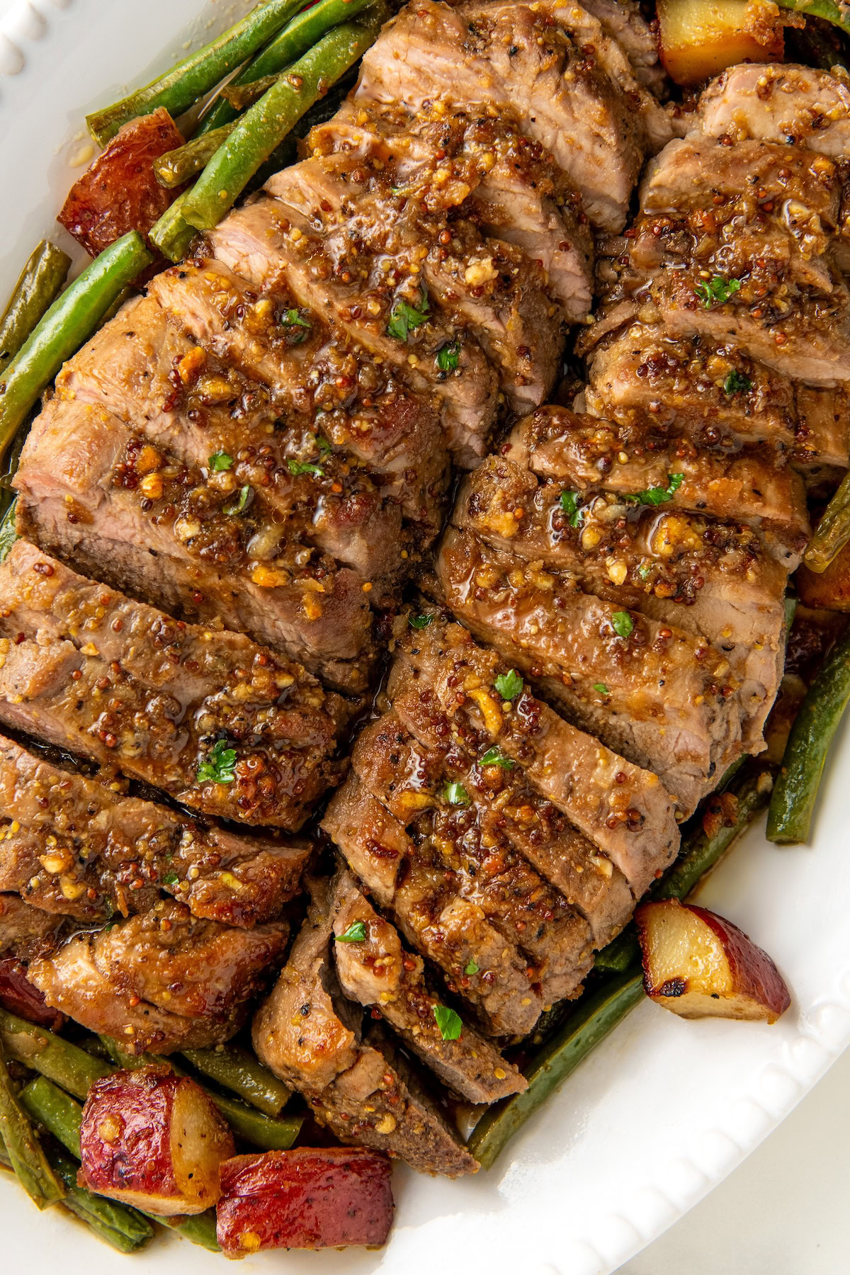 Sliced, honey garlic roasted pork tenderloin served over a bed of green beans and roasted red potatoes on a white serving platter.