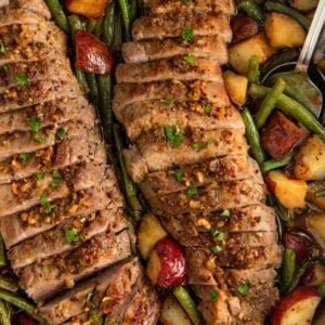 Sliced roasted pork tenderloin with a honey garlic crust, served in a skillet alongside roasted red potatoes and green beans. The dish is garnished with chopped herbs.