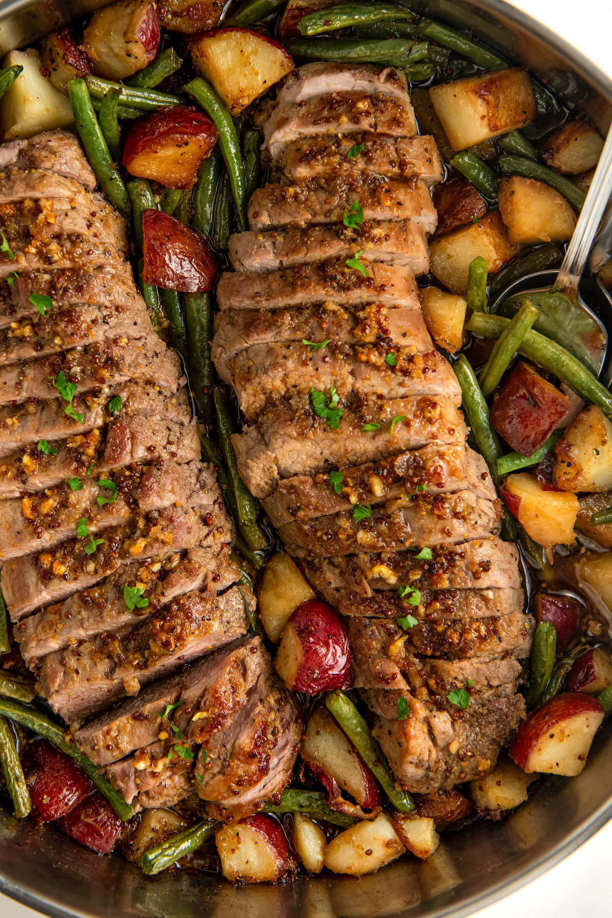 This easy Roasted Pork Tenderloin with Potatoes and Green Beans is packed with bold flavor from a sweet and savory honey garlic glaze. Sliced roasted pork tenderloin with a honey garlic crust, served in a skillet alongside roasted red potatoes and green beans. The dish is garnished with chopped herbs.