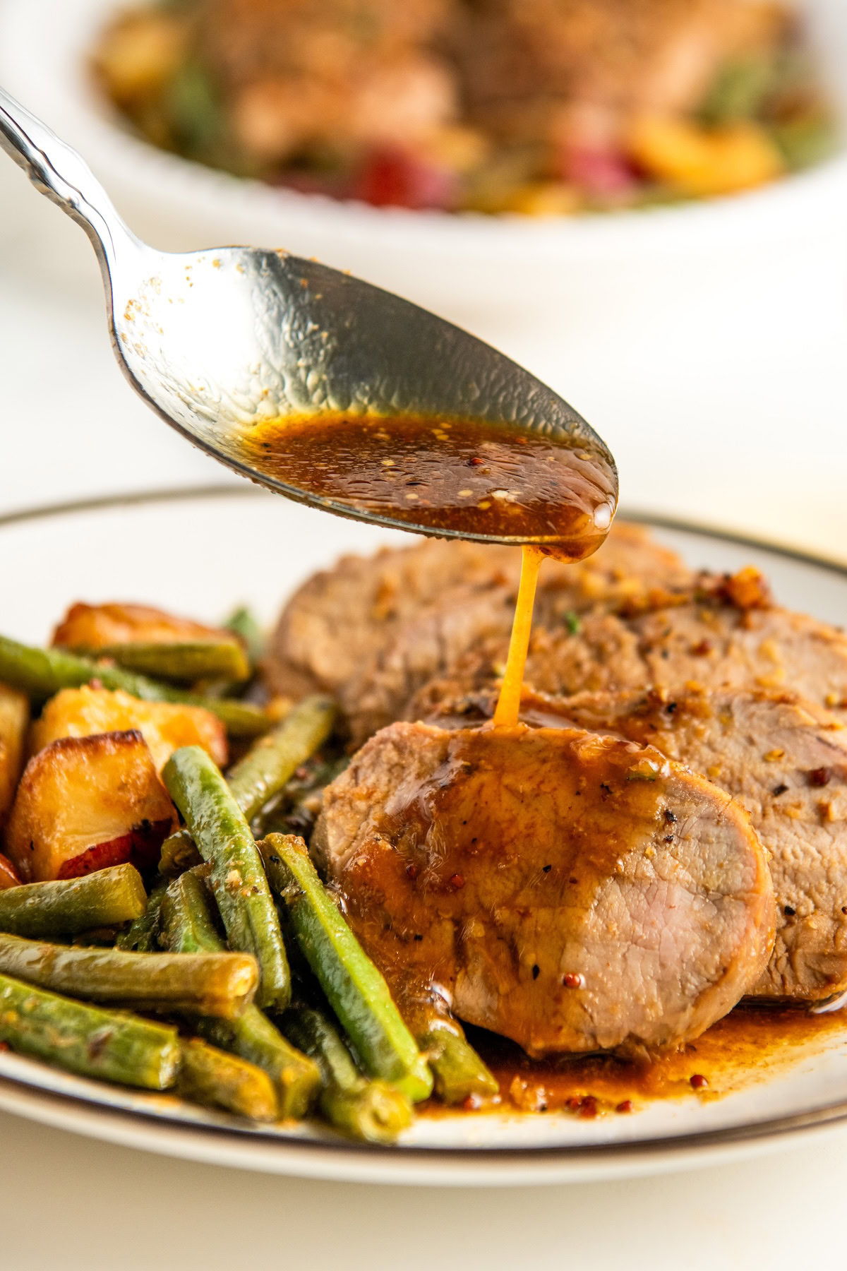 A close-up of a plate with slices of roasted pork tenderloin, green beans, and red potatoes. A spoon is pouring the cooking juices from the skillet over the pork.