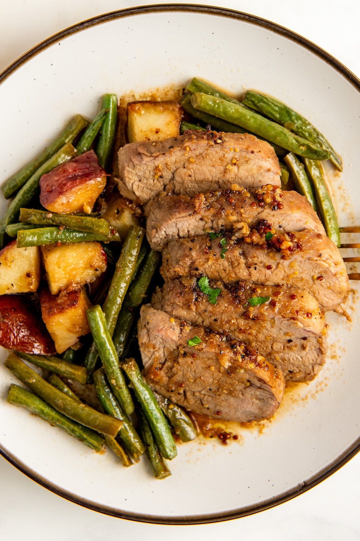 A plate with sliced roasted pork tenderloin topped with fresh herbs, surrounded by cooked green beans and roasted potatoes, all drizzled with a savory honey garlic sauce.