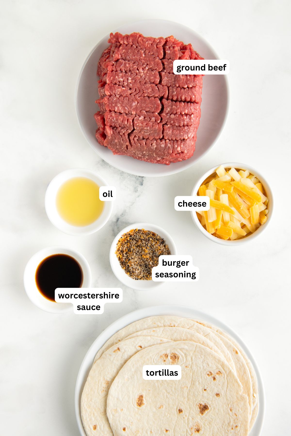 Image of smash burger taco recipe ingredients on a white surface: a plate of raw ground beef, a bowl of shredded cheese, a small bowl of oil, a bowl of burger seasoning, Worcestershire sauce in a bowl, and a plate of tortillas.