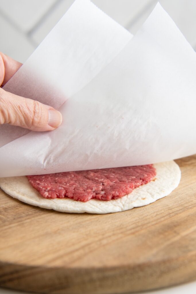A hand lifting parchment paper off a raw burger patty pressed flat on top of a flour tortilla, resting on a piece of parchment on a wooden board.