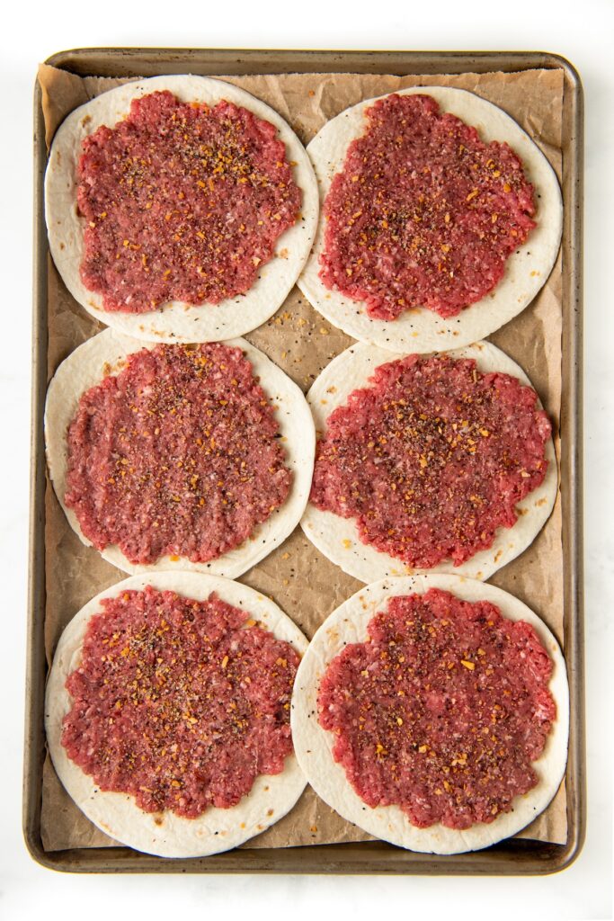 A baking tray with six round flour tortillas topped with raw ground beef mixed with burger seasoning, placed on parchment paper.