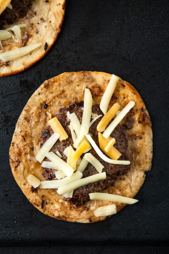 A flour tortilla topped with a seasoned beef patty and a mix of white and yellow shredded cheese, placed on a griddle.