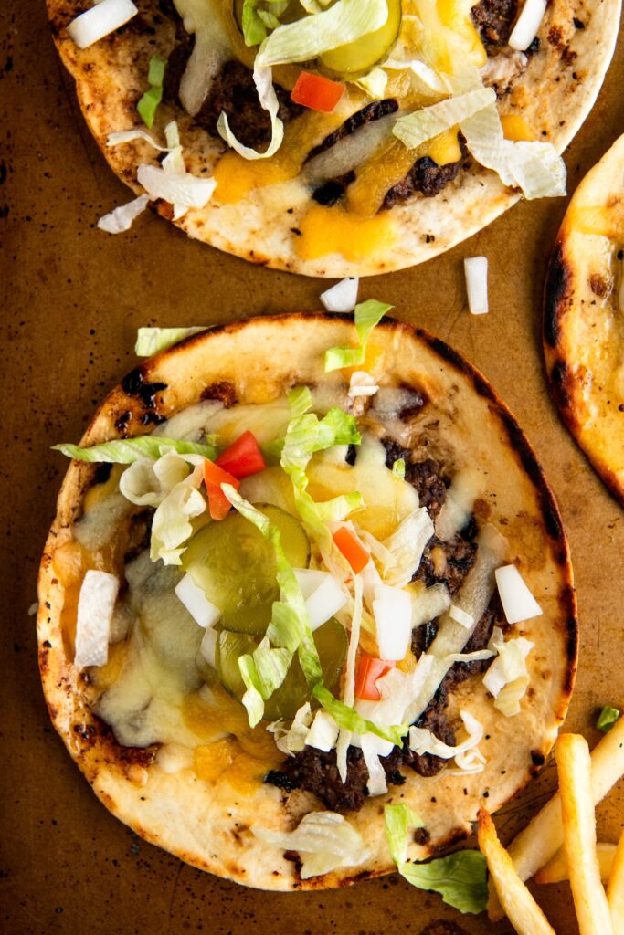 Two smash burger tacos topped with melted cheese, pickles, shredded lettuce, diced tomatoes, and onions on a brown surface. A few French fries are partially visible in the lower part of the image.