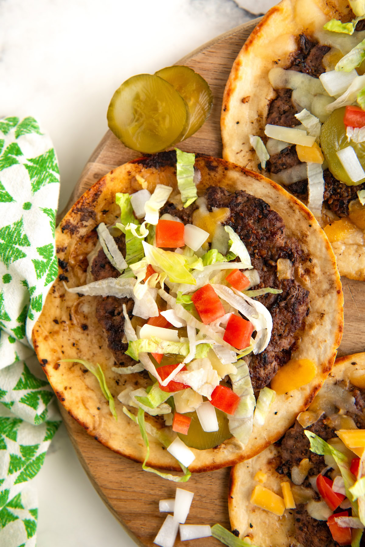 A close-up of Big Mac smash tacos on a wooden board. The tacos are topped with seasoned ground beef, lettuce, diced tomatoes, onions, and burger sauce. Beside the tacos, there are two pickle slices. A green patterned napkin is partially visible.