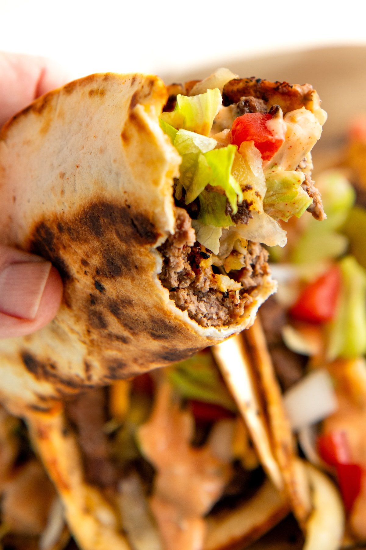 Close-up of a hand holding a smash burger taco with lettuce, diced tomatoes, and burger sauce. The flour tortilla is slightly charred.