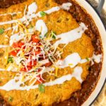 A round baked tamale pie casserole featuring a layer of green chile cornbread on top, covered with melted cheese and a drizzle of sour cream. The dish is garnished with diced tomatoes, shredded cheese, and cilantro, served in a black-handled pot on a striped cloth.