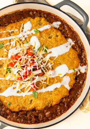 A round baked tamale pie casserole featuring a layer of green chile cornbread on top, covered with melted cheese and a drizzle of sour cream. The dish is garnished with diced tomatoes, shredded cheese, and cilantro, served in a black-handled pot on a striped cloth.