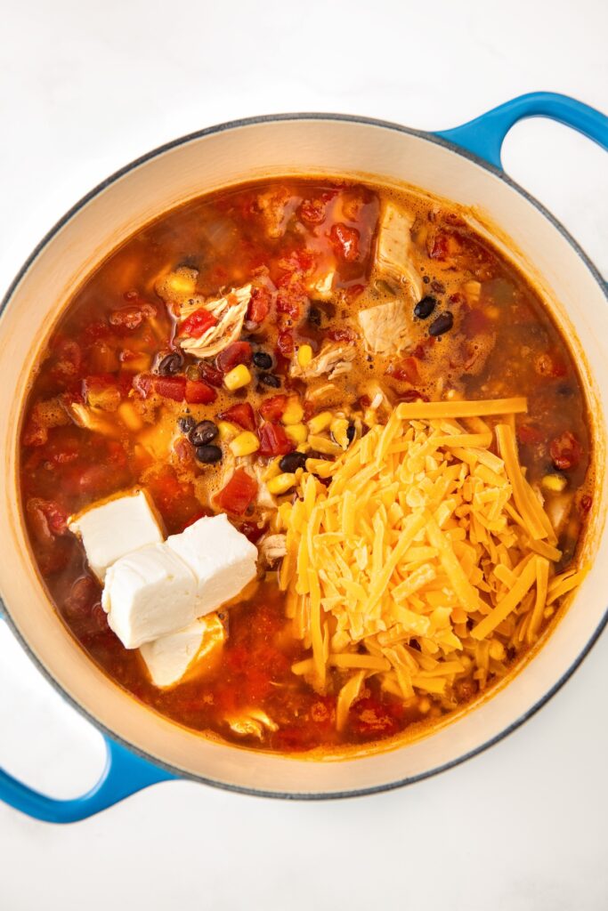 A pot of soup with chunks of chicken, black beans, corn, tomatoes, and a rich broth. Topped with cheddar cheese and cream cheese.