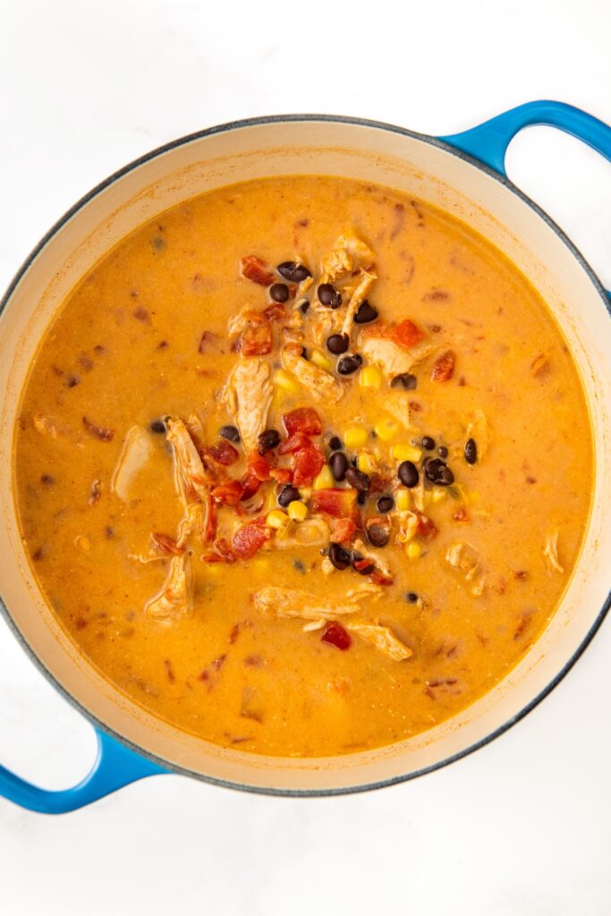 A pot of creamy chicken taco soup with shredded chicken, black beans, corn, and diced tomatoes. The soup has an orange hue and is in a blue-handled pot on a white background.